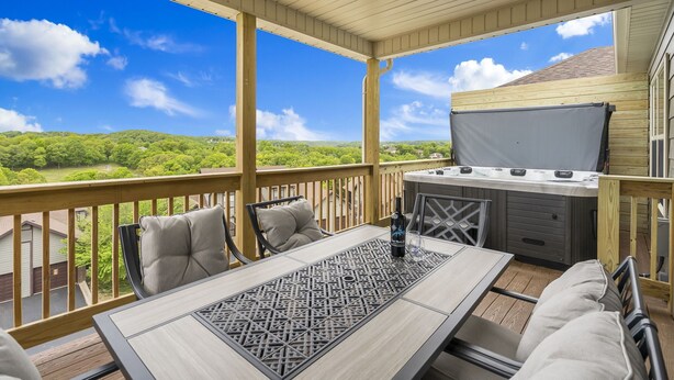 A covered outdoor deck with a table and chairs, a hot tub, and a scenic view of green hills. - Bright Upgraded Decor with Hot Tub, Deck, Arcade, Free Tickets & Onsite Ledgestone Golf (Branson West)