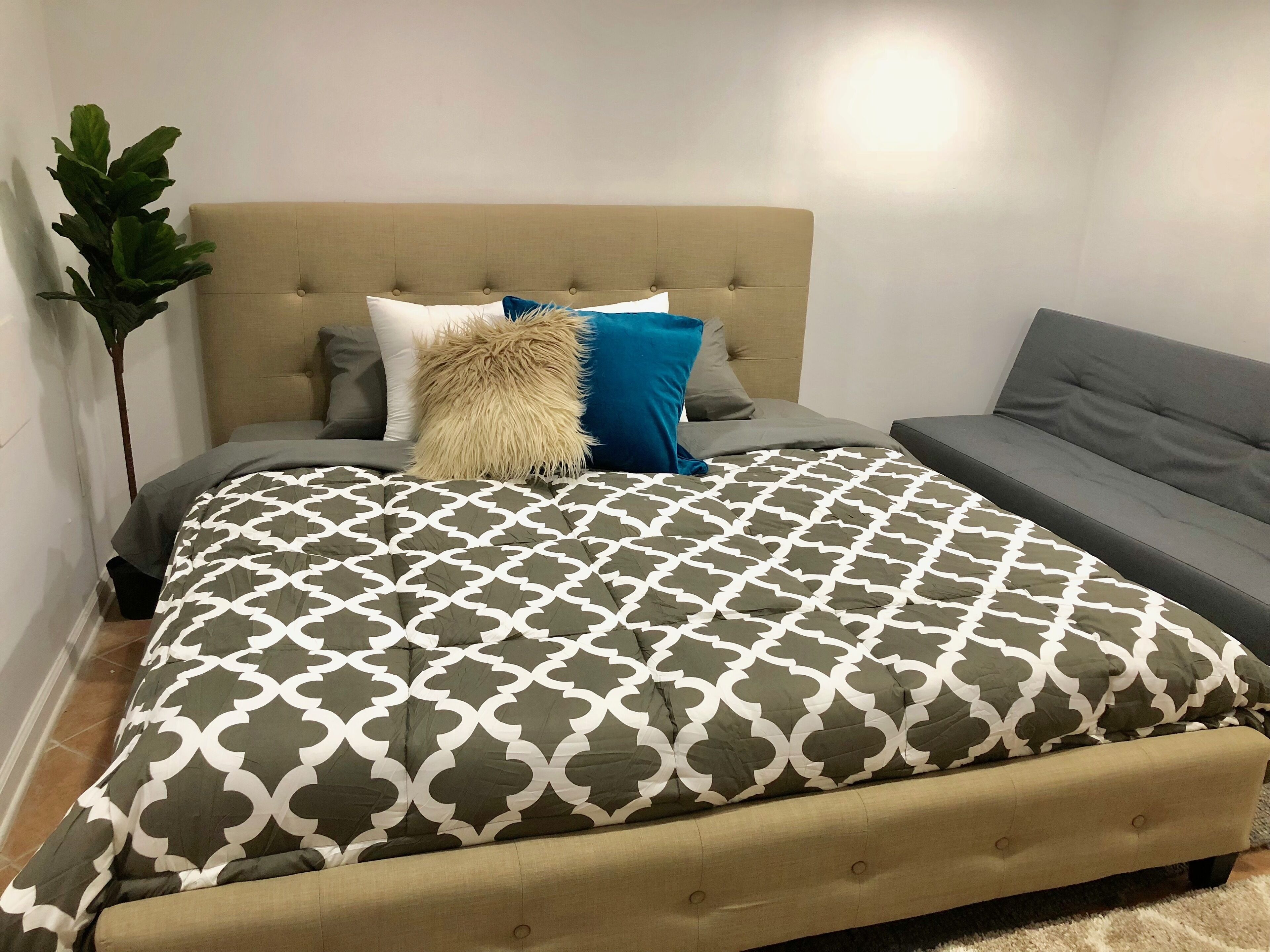 Ett sovrum med en säng, en grå soffa, en stor krukväxt och en beige klädd sänggavel.