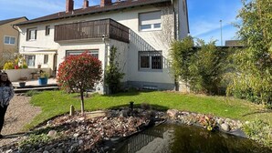 Ein zweistöckiges Haus mit einem Balkon, einem kleinen Teich und einem Garten mit verschiedenen Pflanzen.