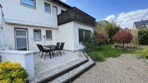 Ein zweistöckiges Gebäude mit einem Balkon, einer Terrasse mit Tisch und Stühlen sowie einem Garten mit verschiedenen Pflanzen.