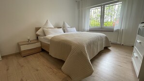 Ein Schlafzimmer mit Bett, Nachttisch, Kommode und einem Fenster mit Blick ins Grüne.