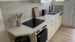 Eine kleine Küche mit eingebautem Herd, Backofen und Spüle. Auf der Arbeitsfläche liegen ein Schneidebrett, ein Wasserkocher und ein Toaster. Die Küche hat eine helle Rückwand und Holzdielen.