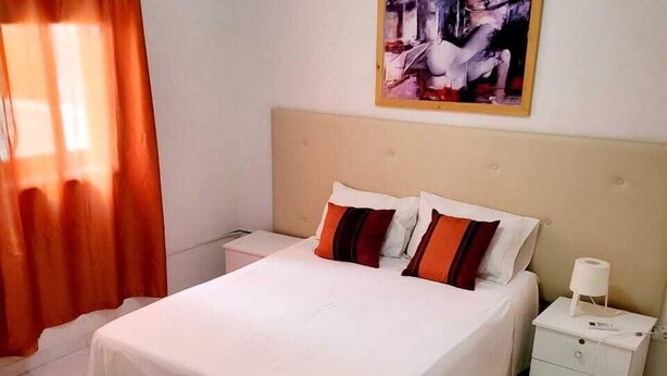 Une chambre d’hôtel avec un grand lit, du linge de lit blanc, une table de chevet, une lampe, un tableau au mur et une fenêtre avec des rideaux.