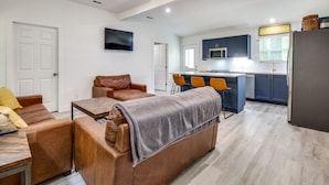 Een moderne woonkamer met een bank, salontafel en een aan de muur bevestigde televisie. Er is een keukenhoek met een magnetron, kastjes en een koelkast.
