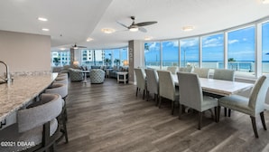 A spacious dining area with a high ceiling, a ceiling fan, and large windows offering a view of the ocean.