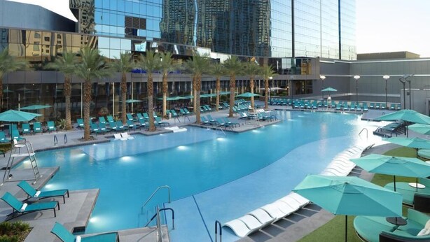 A large indoor swimming pool with multiple lanes, surrounded by lounge chairs and umbrellas, with high-rise buildings in the background. - Stay in Style at Elara Hilton's 2BR Suite During the Las Vegas Formula 1 Race. (Las Vegas)