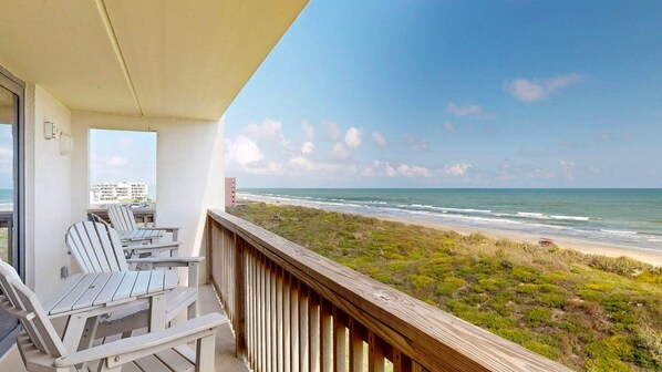 A balcony with white chairs and a wooden railing overlooking a beach with ocean views.