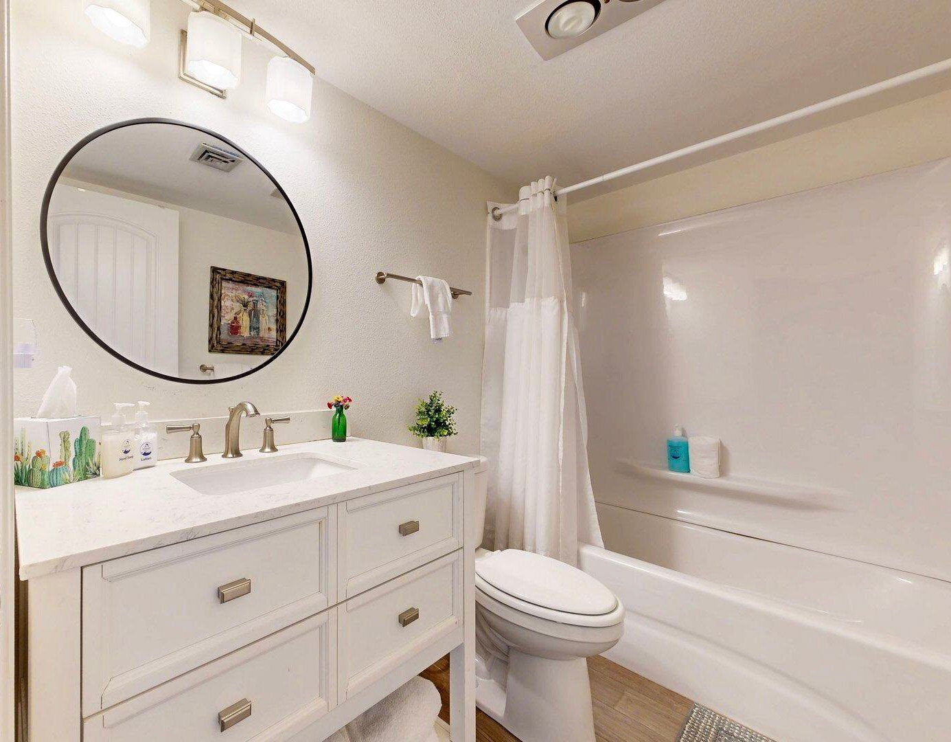 A bathroom with a white vanity, a round mirror, a toilet, and a bathtub.