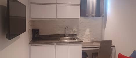 A compact kitchen with white cabinets, a stainless steel sink, and a coffee maker. There is a small trash bin and a chair near the window.