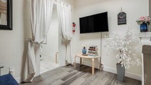A room with a wooden floor, a bed with white curtains, a television mounted on the wall, a small table with books and a vase, and a potted plant. - 4 Beach Flats Sleeps 18 2Blks from Beach,Boardwalk,Sand,Surf (San Diego)