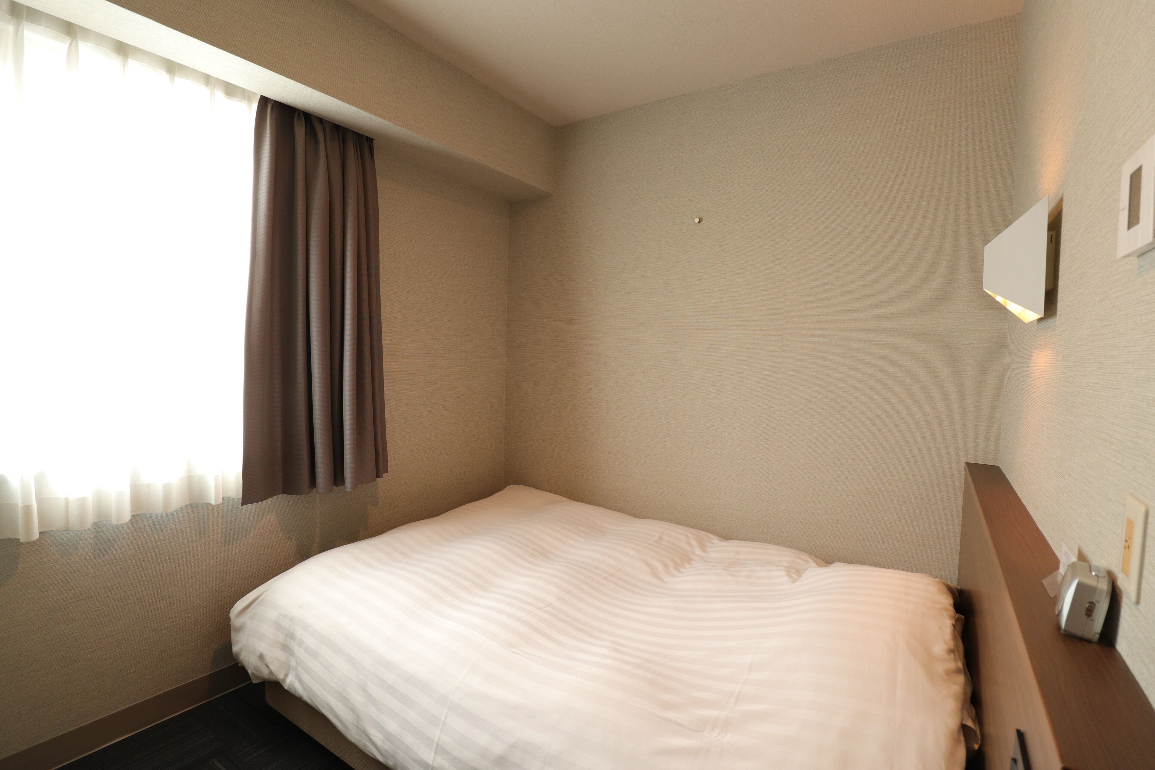 A hotel room with a single bed, a window with curtains, a wall-mounted light, and a small bedside table with a phone and a tissue box.