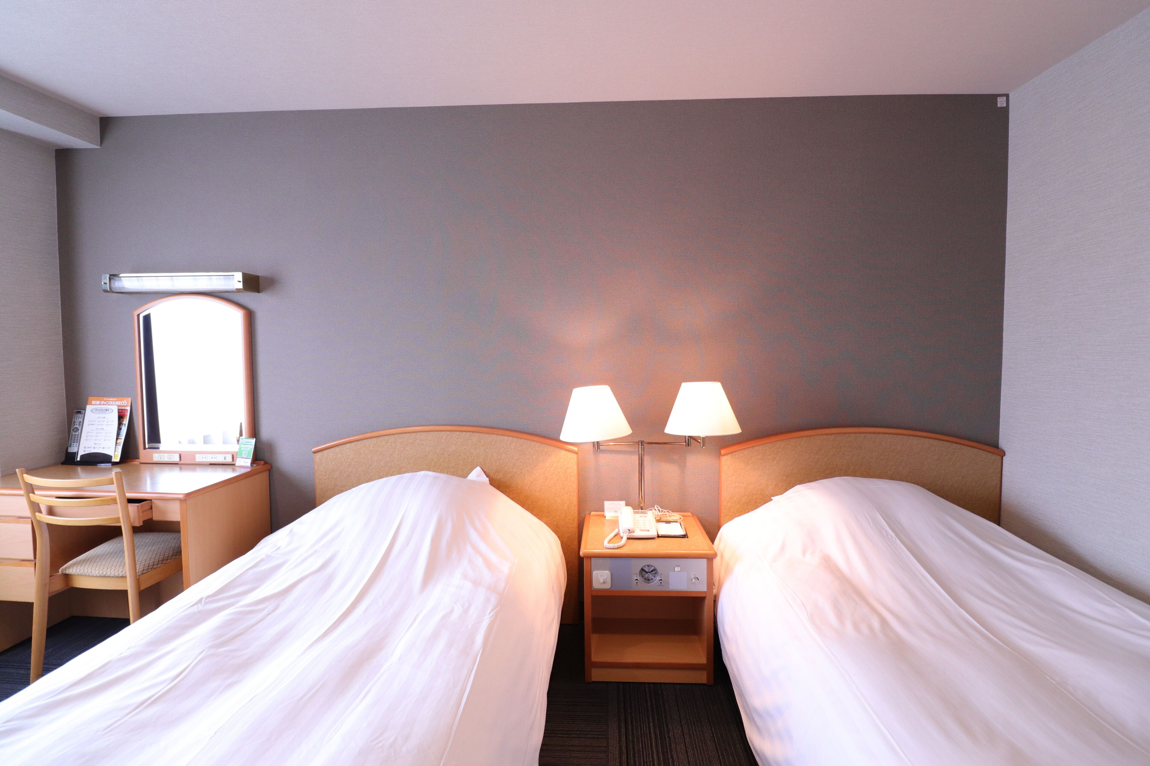 A hotel room with two single beds, a desk with a chair, a nightstand with a lamp, and a wall-mounted mirror.