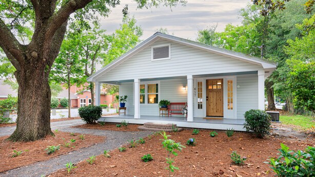 A small, single-story house with a porch, a large tree in front, and a well-maintained garden.