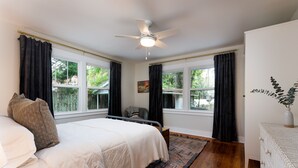 A bedroom with a bed, a desk, a chair, a ceiling fan, and large windows with curtains.