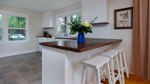 A kitchen with white cabinets, a wooden countertop, and a vase of flowers. There are three white barstools and a framed picture on the wall.