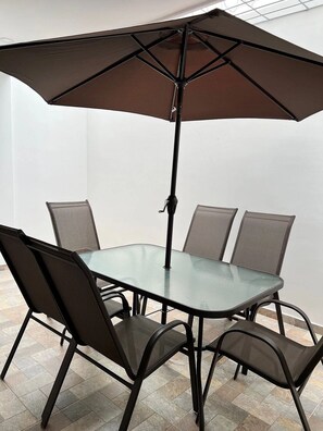 Uma mesa com tampo de vidro, quatro cadeiras, um guarda-sol e estrutura metálica.