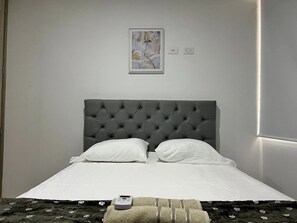 Cama bem arrumada com cabeceira estofada, duas almofadas brancas e um controle remoto sobre uma mesinha.