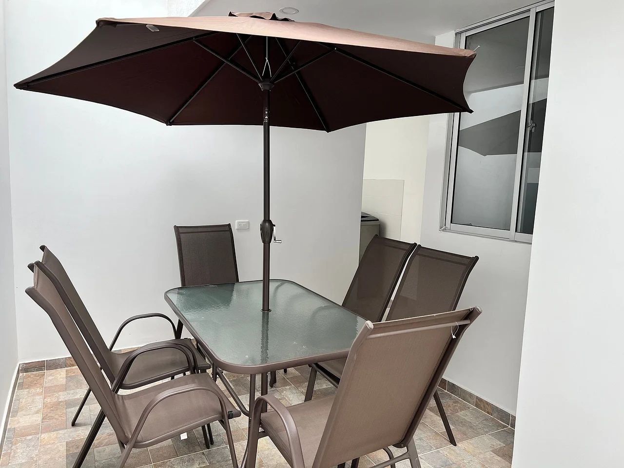 Un comedor con mesa de vidrio, sillas metálicas, una sombrilla grande y una ventana con persianas.