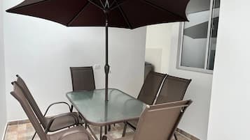 Un comedor con mesa de vidrio, sillas metálicas, una sombrilla grande y una ventana con persianas.