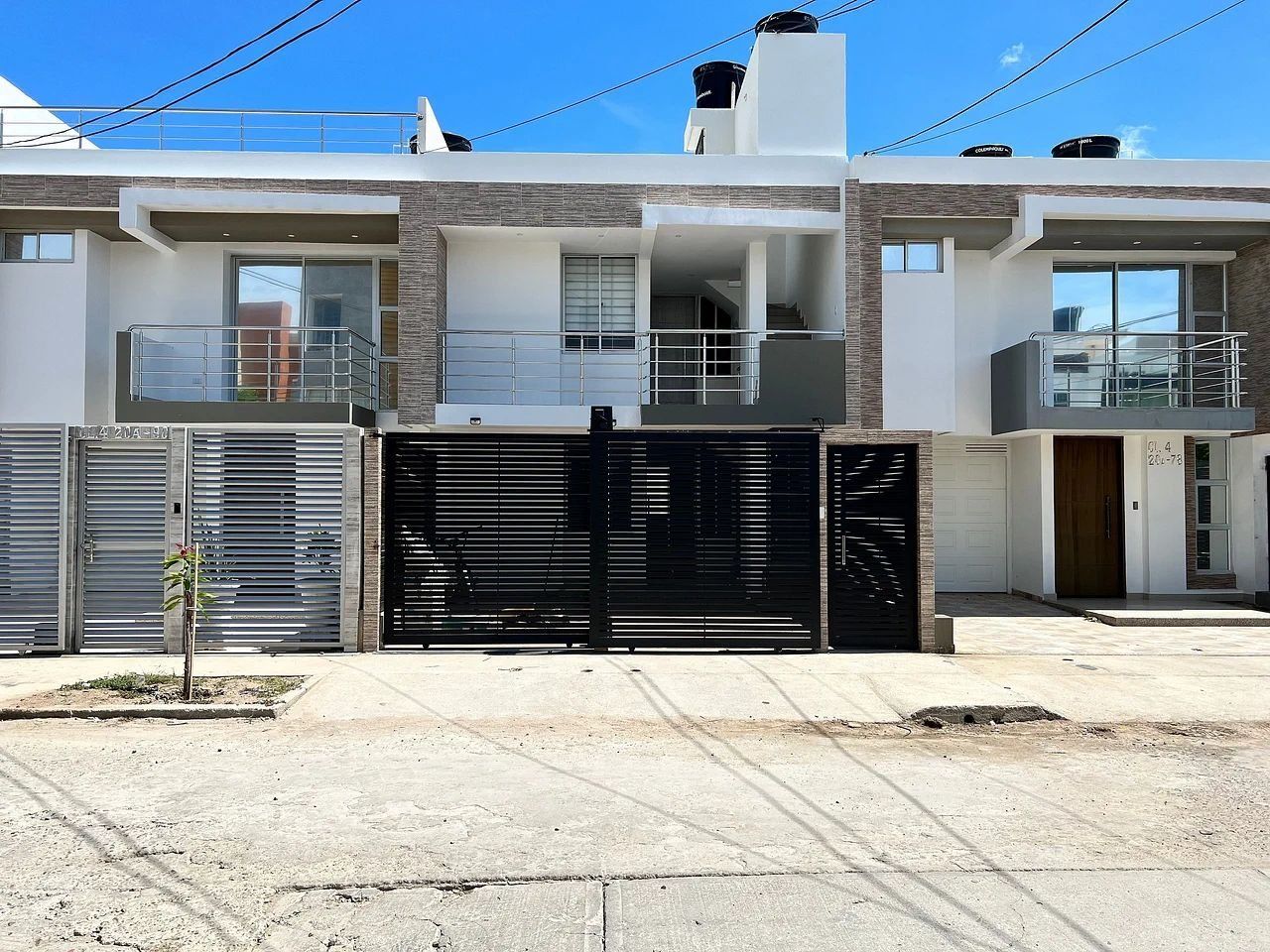 Un edificio residencial moderno de dos pisos, con entrada protegida por una reja, un pequeño jardín y cielo despejado.