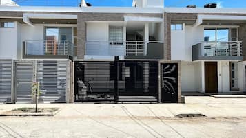 Un edificio residencial moderno de dos pisos, con entrada protegida por una reja, un pequeño jardín y cielo despejado.