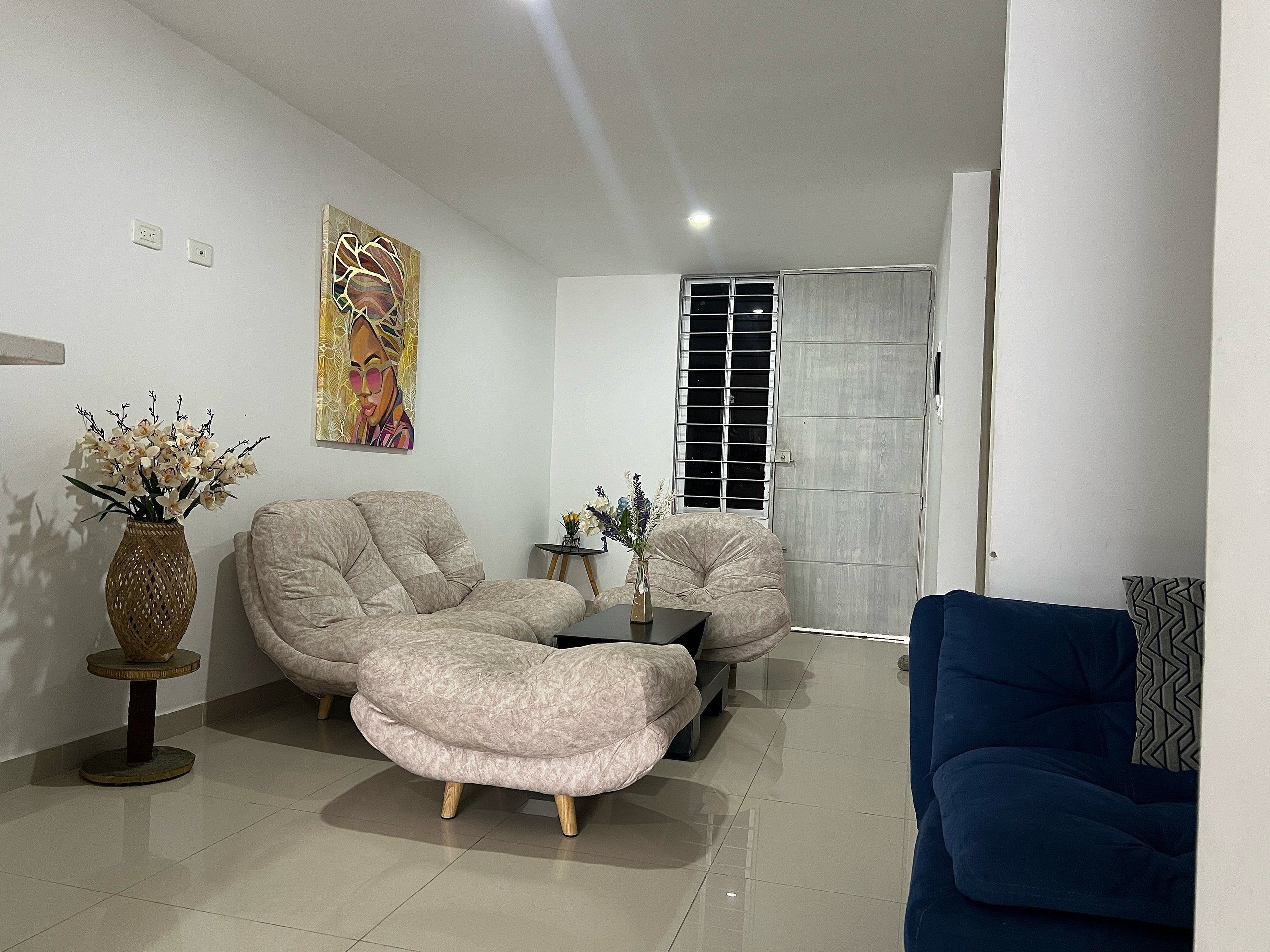 A living room with a sofa, a coffee table, and a blue armchair. There is a painting on the wall and a window with a metal grid.