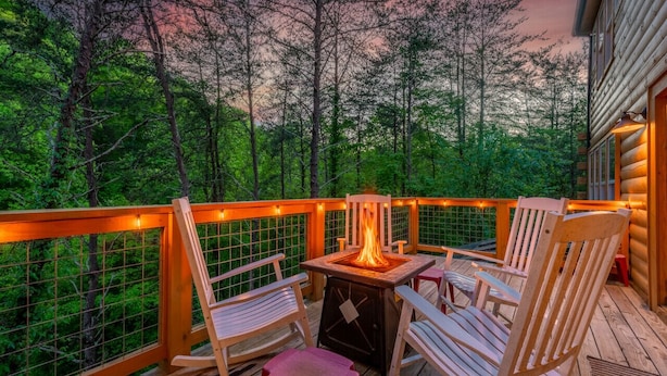 Eine Holzterrasse mit Feuerstelle und Stühlen, umgeben von einem Hollerleger und Blick auf ein bewaldetes Gebiet.