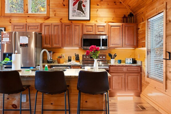 Cocina de cabaña de madera con un refrigerador de acero inoxidable, microondas y estufa. Hay armarios de madera, un fregadero y una ventana con persianas.