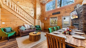 A wooden cabin interior with a dining area, a sofa set, a staircase, and a stone fireplace. - Heavenly Heights by Ghosal Luxury Lodging (Sevierville)