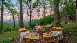 A cozy outdoor seating area with a stone fire pit surrounded by wooden tables and benches, set against a backdrop of trees and a distant view.
