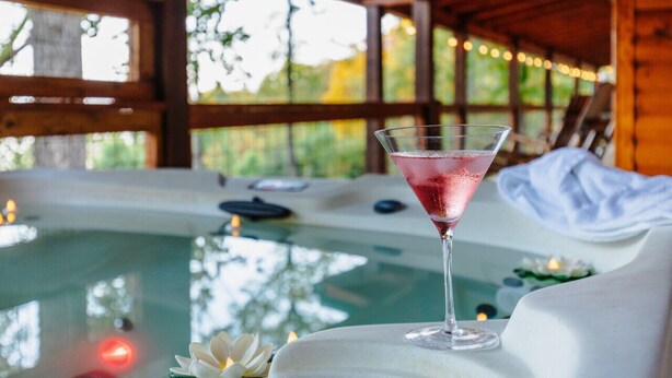 A clear glass of pink drink on a table with a view of a hot tub and outdoor greenery.