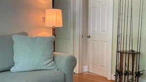 Un salon avec un canapé gris, une table basse en bois sur laquelle repose un panier en osier, une lampe, et une porte derrière laquelle on aperçoit des cannes à pêche.