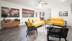 A modern living room with a yellow sofa, black chairs, a wooden cabinet, and a ceiling fan.