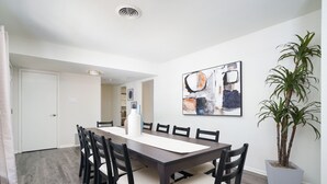 A dining area with a long table, chairs, a potted plant, and artwork on the wall.