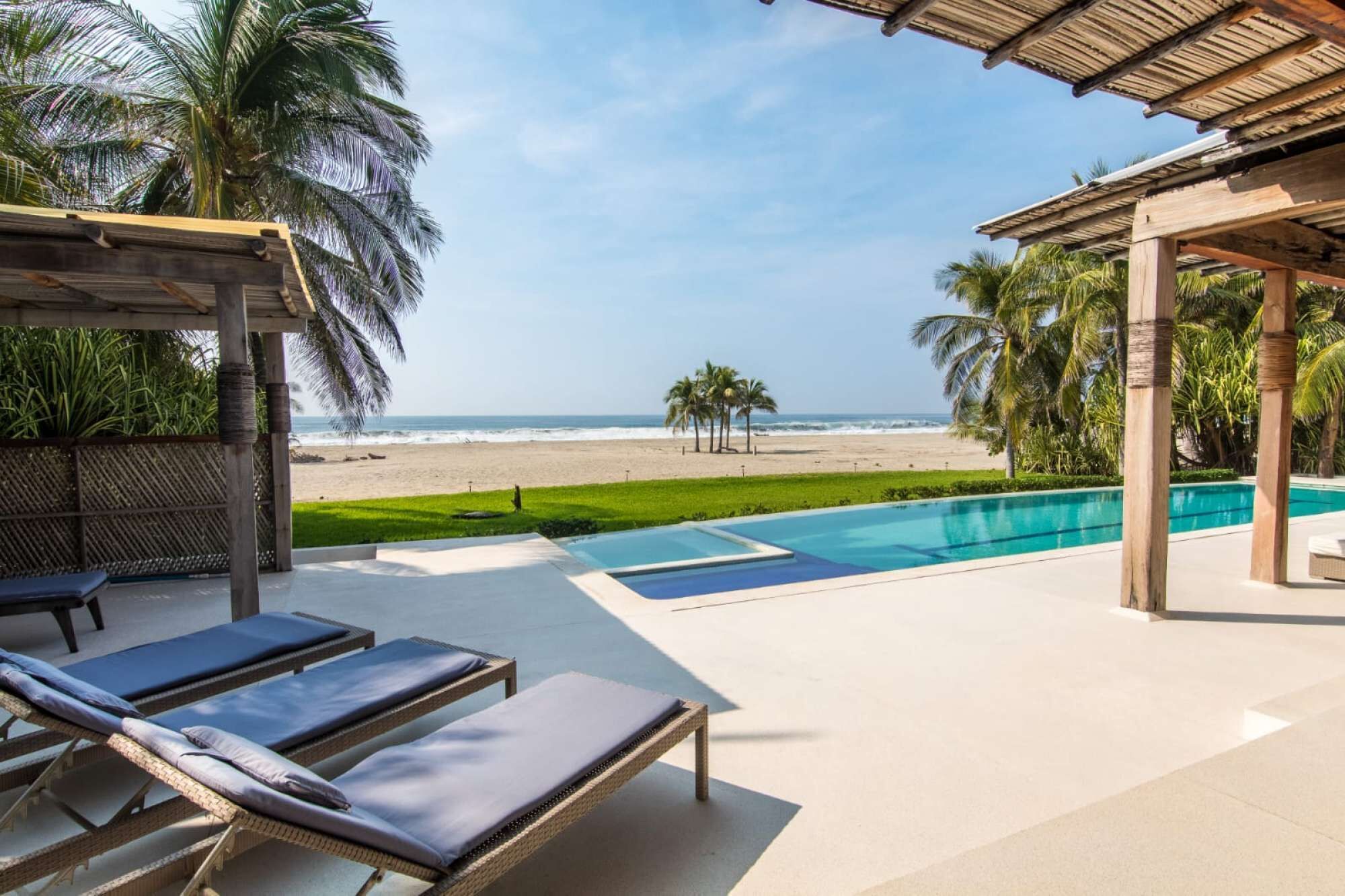 Zona de piscina con sillones, vista a la playa y palmeras.