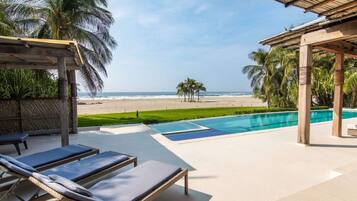 Zona de piscina con sillones, vista a la playa y palmeras.
