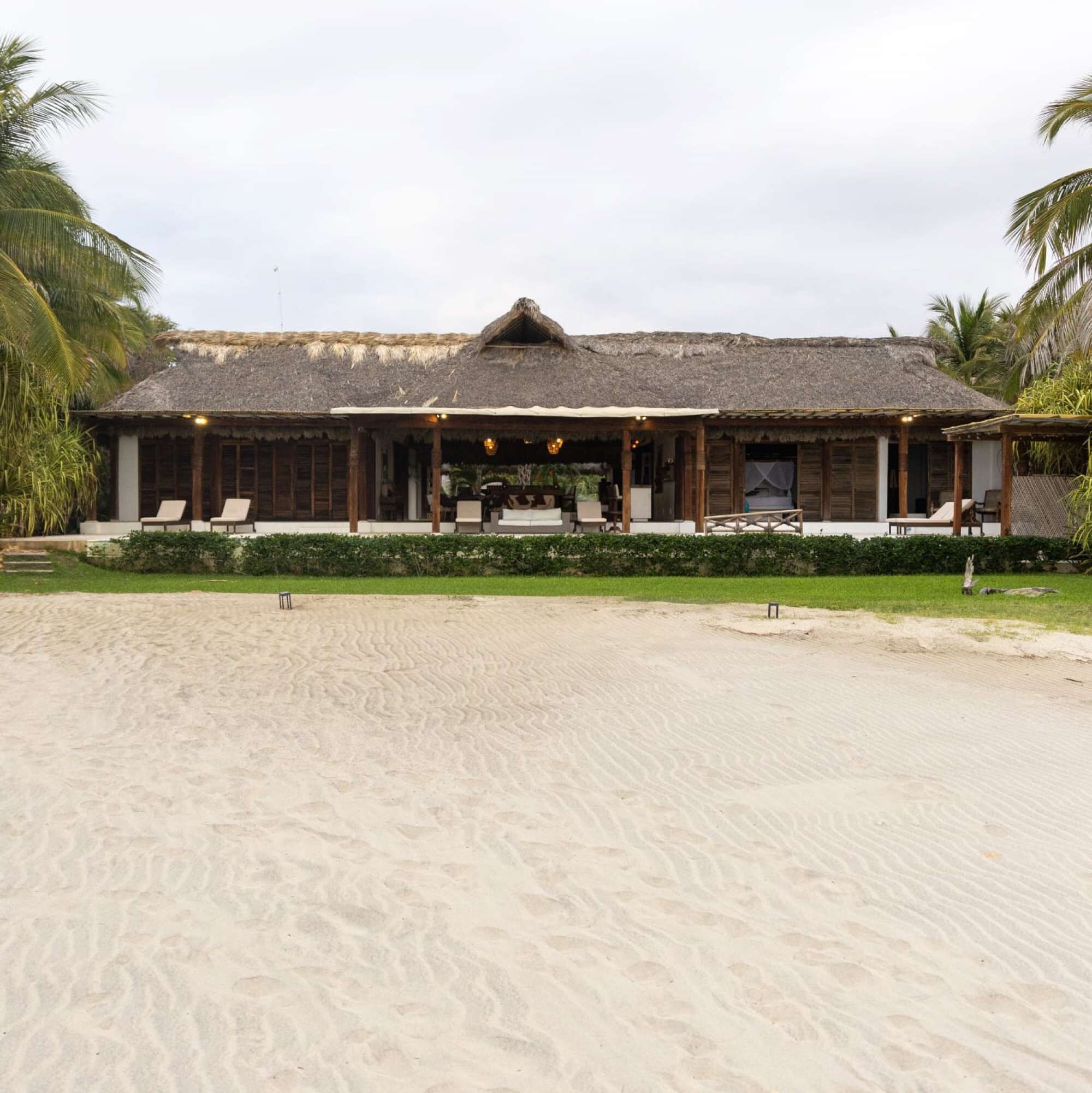 Una propiedad frente a la playa con un edificio de techo de paja, mobiliario exterior y una playa de arena.