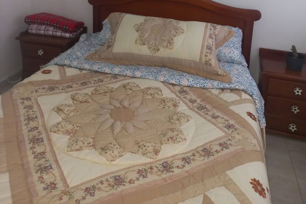 A bedroom with a wooden bed, a nightstand with a soccer ball drawer, a small shelf with a bottle, and a red plaid blanket on the nightstand.