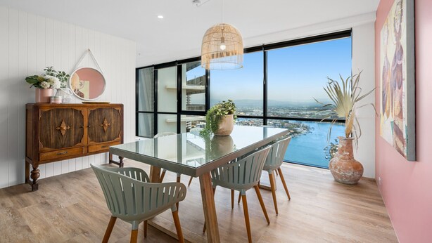 Un comedor moderno con una mesa de vidrio, sillas de madera y vistas a un paisaje urbano a través de amplios ventanales.