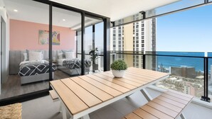 Un balcón con una mesa y sillas de madera, una planta en maceta y vistas al océano y edificios.