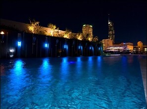 Une piscine nocturne avec des jeux d’eau illuminés et une vue sur la ville en arrière-plan.