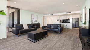 Ein modernes Wohnzimmer mit einer Couch und einem Couchtisch, ein Küchenbereich mit Geräten und eine Glastür, die in einen anderen Raum führt.