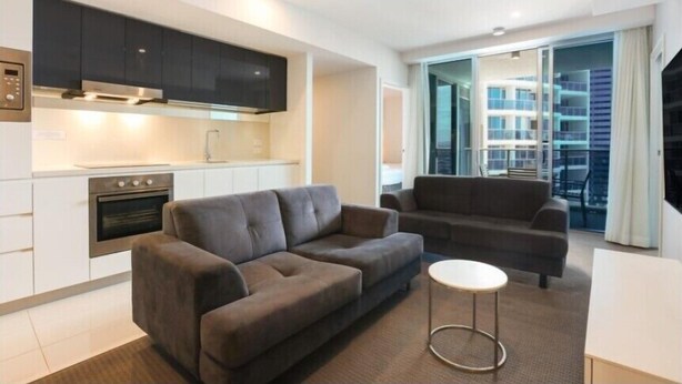 A modern apartment with a kitchen, two sofas, a small round table, and a balcony with a city view. - 26 levels above Hilton hotel - 2 bedrooms (Surfers Paradise)