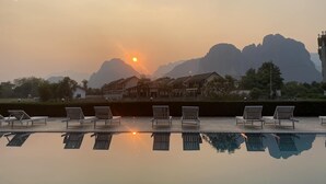 A swimming pool with lounge chairs, a sunset, and mountains in the background. - Lisha Grand Riverside Hotel (Vang Vieng)