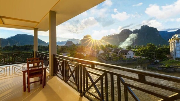 A balcony with a wooden chair, overlooking a river with mountains in the background and a sunrise. - Lisha Grand Riverside Hotel (Vang Vieng)