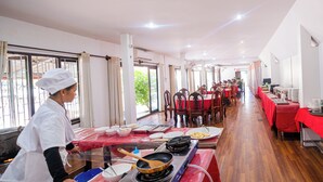 A spacious dining area with multiple tables covered in red tablecloths, a chef preparing food, and large windows allowing natural light. - Lisha Grand Riverside Hotel (Vang Vieng)