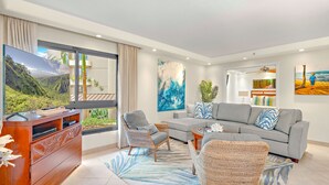 A modern living room with a large TV, grey sofa, wicker chairs, and a view of a mountainous landscape through a sliding door.
