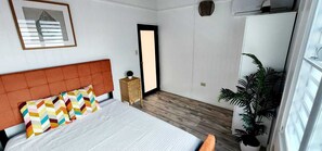 Un dormitorio con cama, cabecera, mesita de noche con lámpara, puerta y planta en maceta.