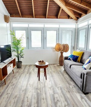 Una sala de estar con techo de madera, un sofá gris, un mueble para TV, una planta en maceta y una mesa redonda de madera.