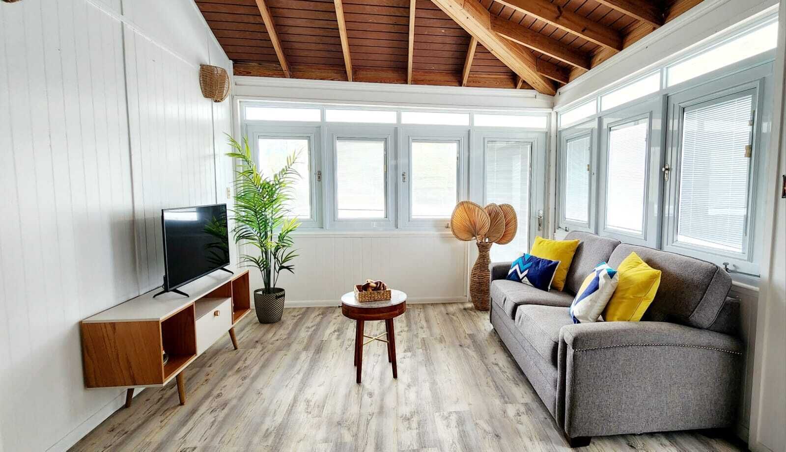Un salon avec un plafond en bois, un canapé gris agrémenté de coussins colorés, un téléviseur posé sur un meuble en bois, une plante en pot et une lampe en osier.